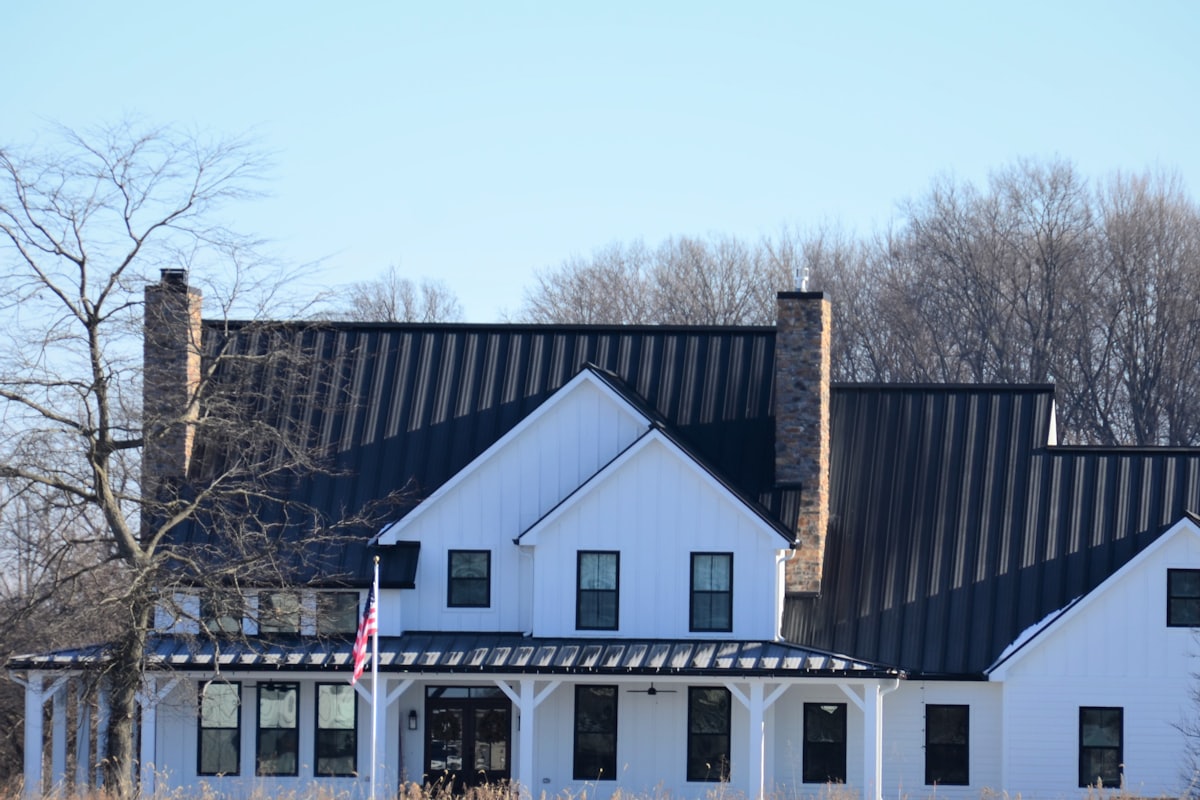 A white board and batten farmhouse with a black standing seam metal roof and two stone chimneys